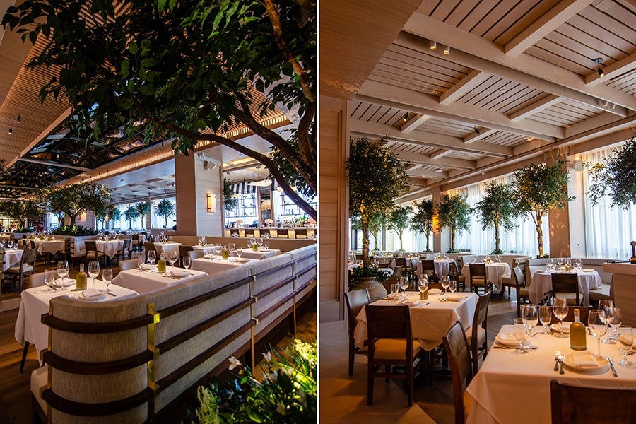 Upscale restaurant interior with neatly arranged tables covered in white tablecloths, set with wine glasses, plates, and cutlery. The space features wooden beams on the ceiling, large windows with sheer curtains, and numerous potted trees adding greenery throughout the dining area. The atmosphere is warm and inviting with soft lighting.