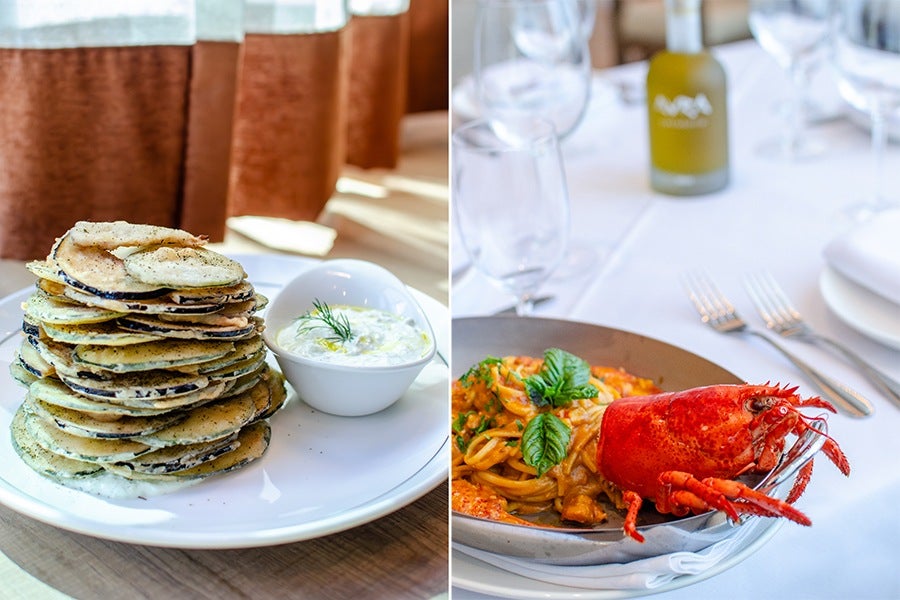 Stack of thin, round pancakes with visible herbs or seasoning, served on a white plate with a small bowl of creamy white sauce garnished with a sprig of dill. Next to it, a dish of pasta with tomato-based sauce, topped with fresh basil leaves and a whole cooked lobster, presented on a gray plate with a white tablecloth and cutlery in the background.