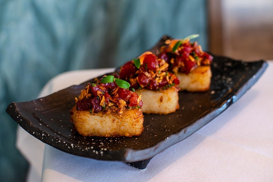 Three pieces of crispy rice topped with diced raw tuna, garnished with small green leaves and crispy fried onions, served on a black rectangular plate.