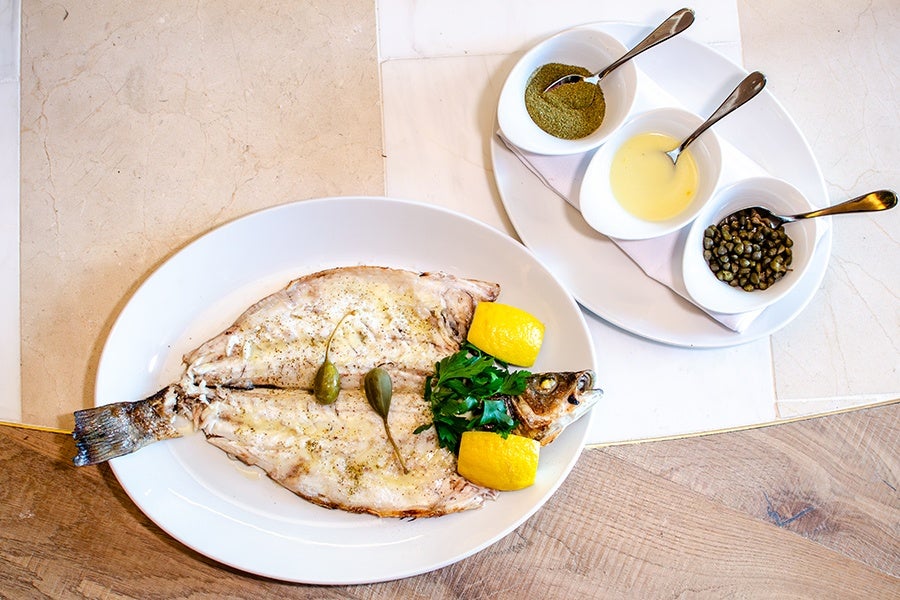 Grilled whole fish served on a white oval plate with two lemon wedges and fresh parsley, accompanied by three small white bowls containing green seasoning, a light yellow sauce, and capers, each with a spoon, on a separate white plate.