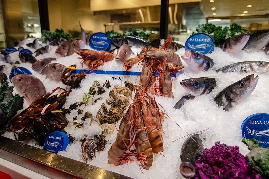 Fresh seafood displayed on a bed of crushed ice, including whole fish, large prawns, lobsters, and various shellfish. The seafood is arranged neatly with some green leafy garnishes and blue labels indicating the types or sources of the seafood. The setting appears to be a market or seafood counter with a stainless steel edge.