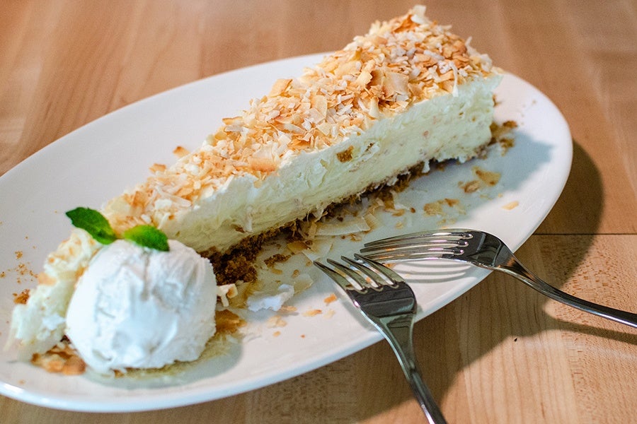 A slice of creamy coconut pie topped with toasted coconut flakes on a white plate, served with a scoop of vanilla ice cream garnished with a small mint leaf. Two forks rest on the plate beside the pie. The plate is on a wooden surface.