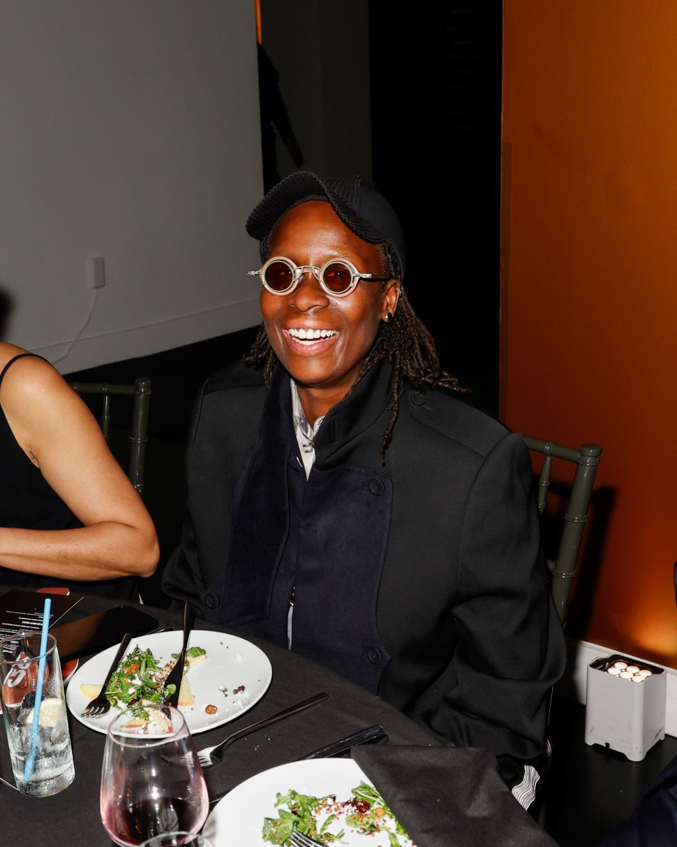 A guest in a black outfit and round tinted glasses smiling while seated at a dinner table with partially eaten plates.