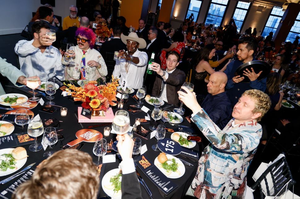 A large group of gala attendees seated around a round dinner table raising glasses in a toast, with plates of food and floral centerpieces.