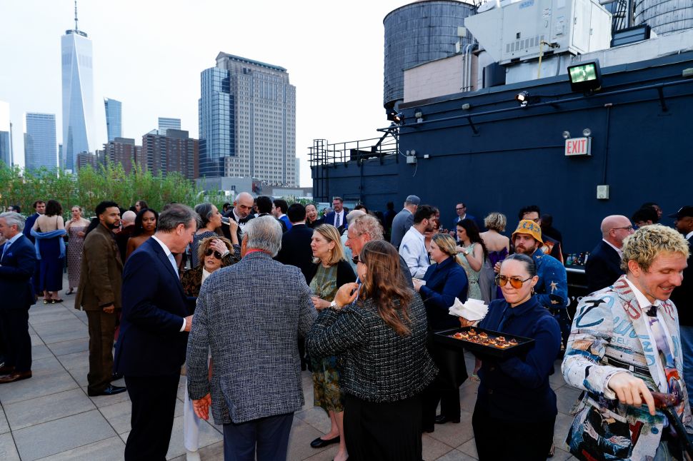 A crowd of guests mingling on a rooftop terrace at sunset with the New York City skyline, including One World Trade Center, in the background.