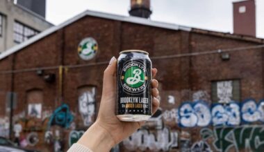 A hand holds a can of Brooklyn Lager in front of the brick brewery .