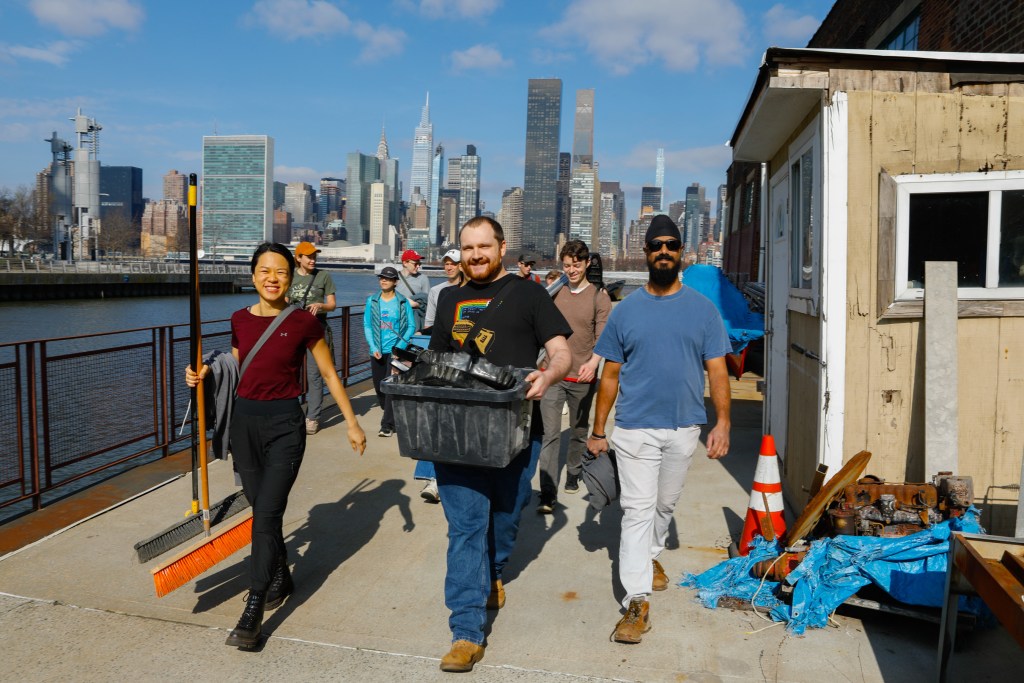 Smiling volunteers hold brooms and cleaning supplies as they walk down Anable Basin in Long Island City. The sun is shining and Manhattan skyline can be seen across the East River.
