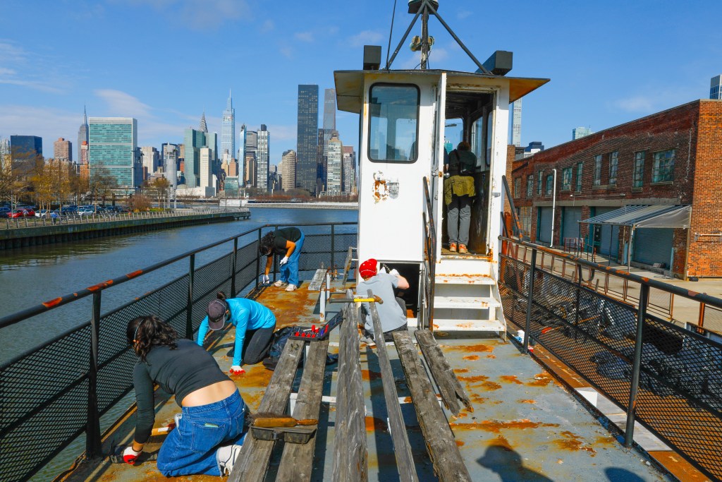 Volunteers scrape paint off the boat deck. The sun is shining and Manhattan skyline can be seen across the East River.