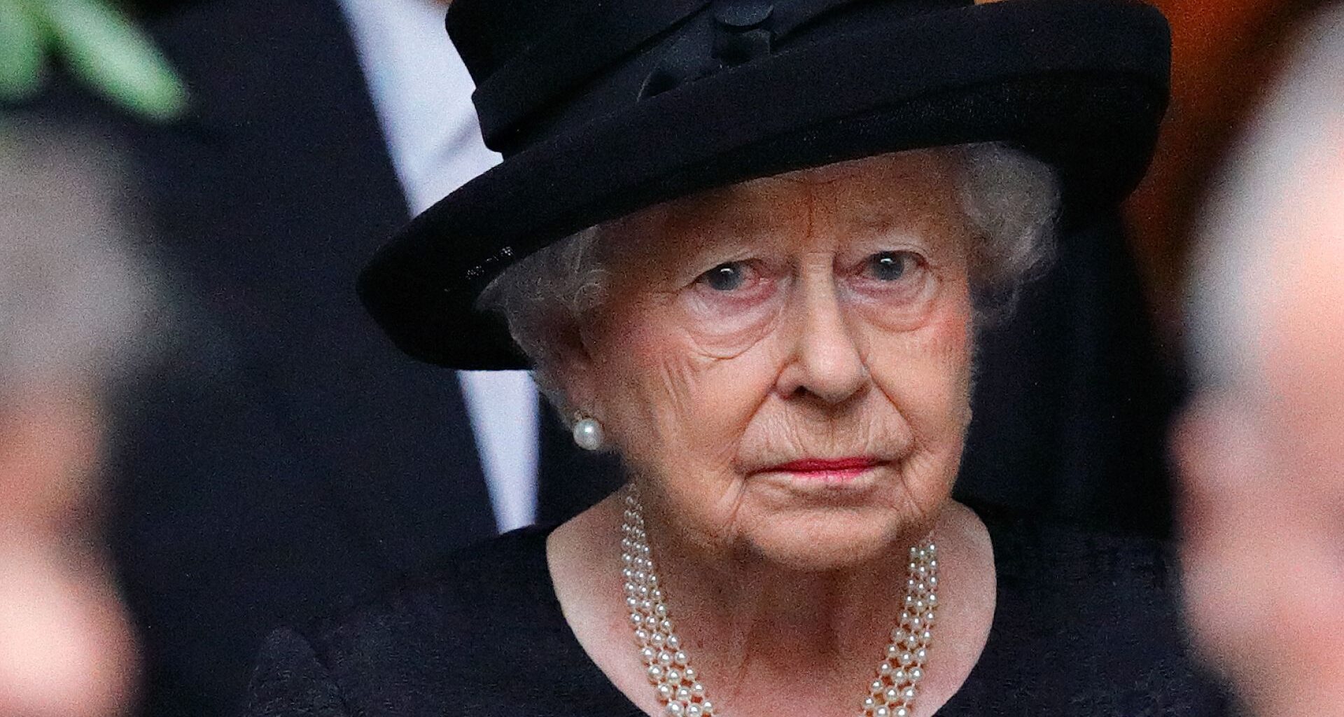 Queen Elizabeth II wears a black dress and matching hat and looks very sad while wearing a string of pearls