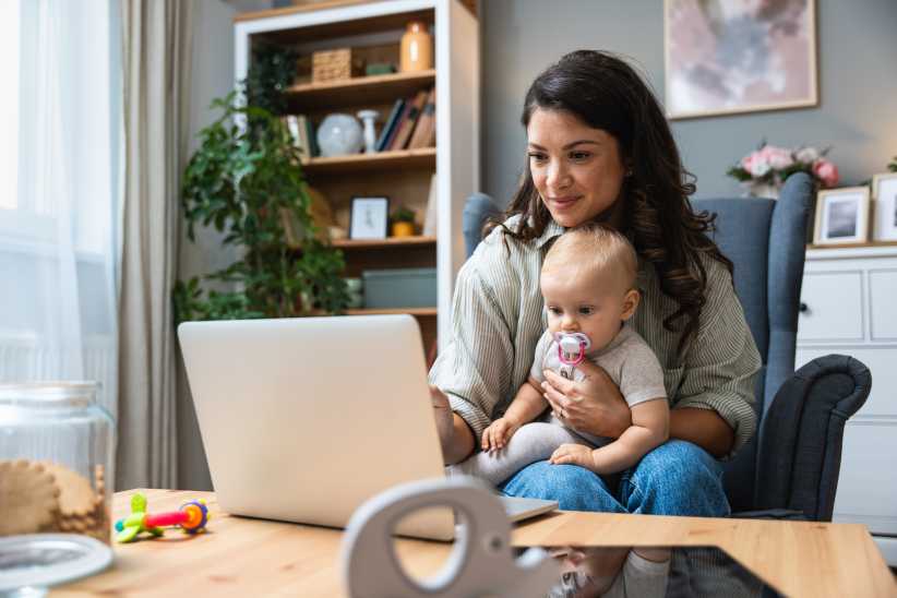 Mom working on laptop while taking care of her baby. New NYC Website Helps Parents Find Childcare — Including Free and Low-Cost Options