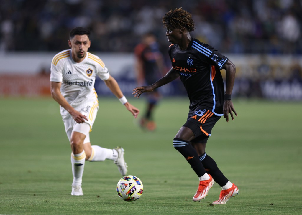 A New York City FC player in a black and blue uniform dribbling the ball while a Los Angeles Galaxy player in a white and gold uniform pursues him.