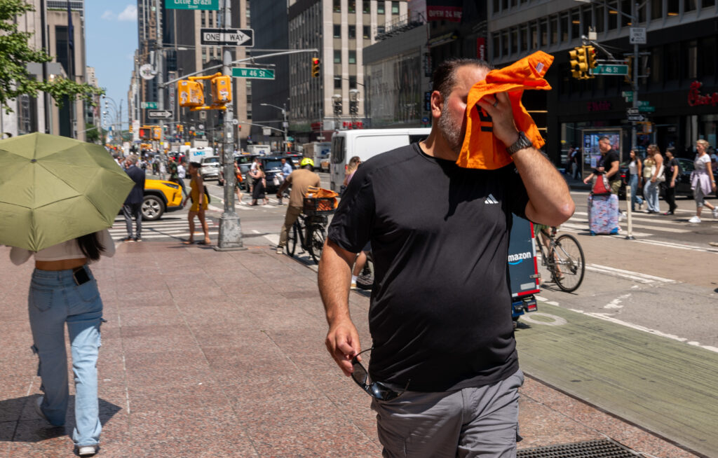 A man on the street wipes his face with orange fabric and another pedestrian holds an umbrella to block out the sun.