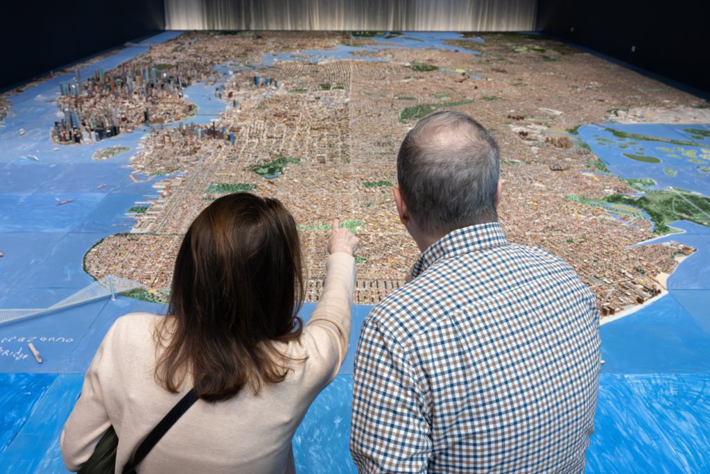 Two adults seen from behind pointing at a large-scale miniature model of New York City spread across a gallery floor.