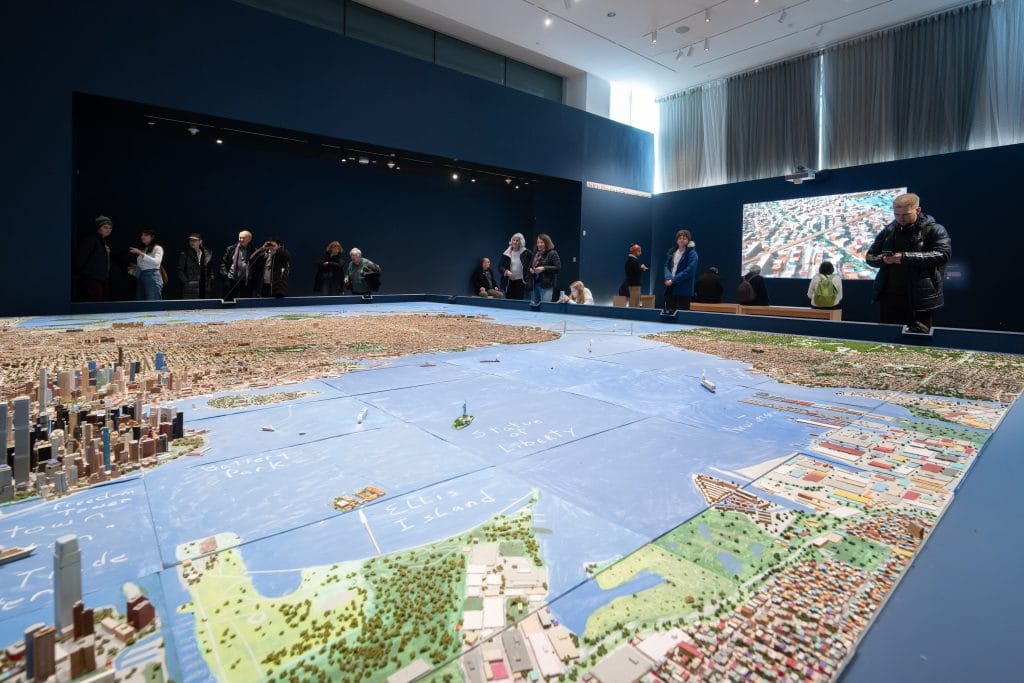 Museum visitors gathered around an expansive city model installation showing New York Harbor, bridges, and boroughs.