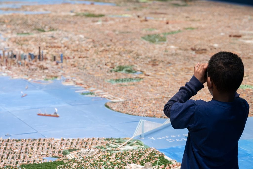 Museum visitors gathered around an expansive city model installation showing New York Harbor, bridges, and boroughs.