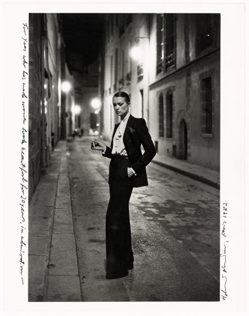 Black-and-white photograph of woman in tailored suit standing on empty nighttime street, holding cigarette amid moody atmosphere
