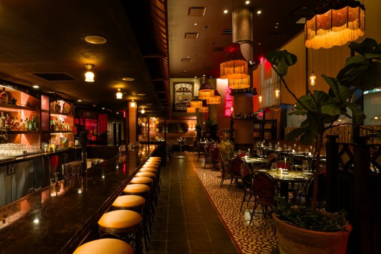 Dimly lit Asian restaurant interior with long bar, fringe pendant lights, red neon Chinese signage, and candlelit dining tables.