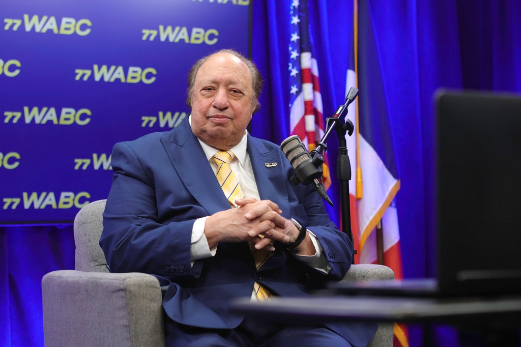 A man in a blue suit and striped yellow tie sitting in a gray armchair, in front of a blue background with the "77 WABC" logo.