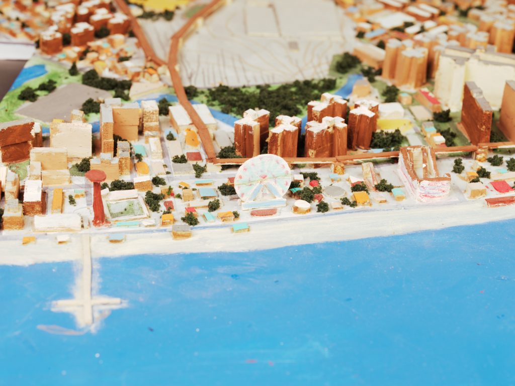 Close-up of a small-scale coastal neighborhood model with buildings, greenery, and a Ferris wheel near the water.