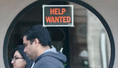 Pedestrians walk past a help wanted sign posted on the door of a restaurant