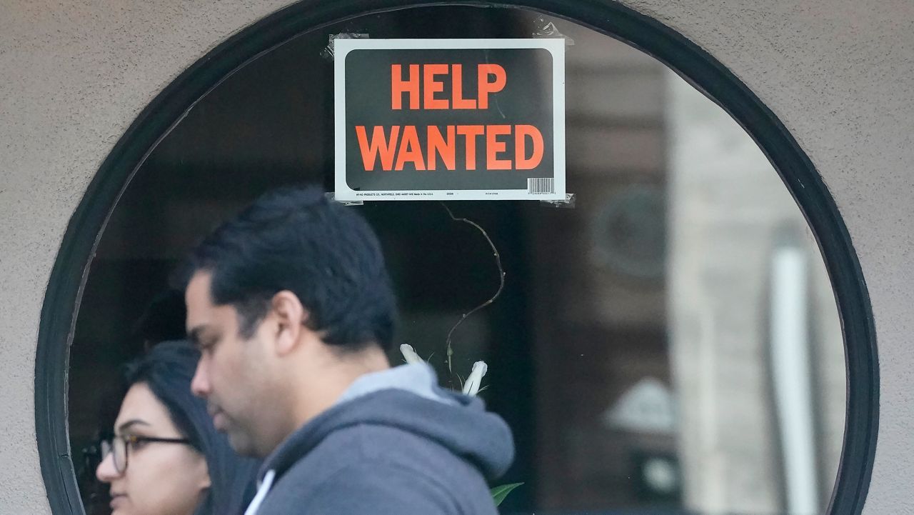 Pedestrians walk past a help wanted sign posted on the door of a restaurant