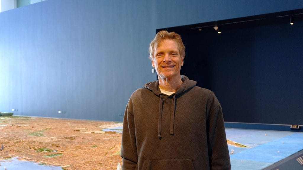 a casually dressed man standing inside a large, modern gallery or museum space. He’s wearing a dark hoodie and smiling at the camera, giving a relaxed, approachable vibe. Behind him is a very large installation or model spread across the floor—it appears detailed and textured, possibly representing a landscape, terrain, or city-like layout. The walls are tall and minimal, with a dark accent wall on the right, which makes the space feel expansive and contemporary. The lighting is soft and even, highlighting both the person and the installation without harsh shadows, suggesting a curated exhibition setting.