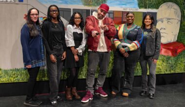 A group of art students, their professor, and The Kid Hero pose in front of a mural