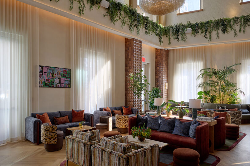 Modern lounge by LEGEARD STUDIO with assorted sofas, patterned armchairs, indoor plants, large windows with sheer curtains, and greenery along a high ledge, all illuminated by natural light.