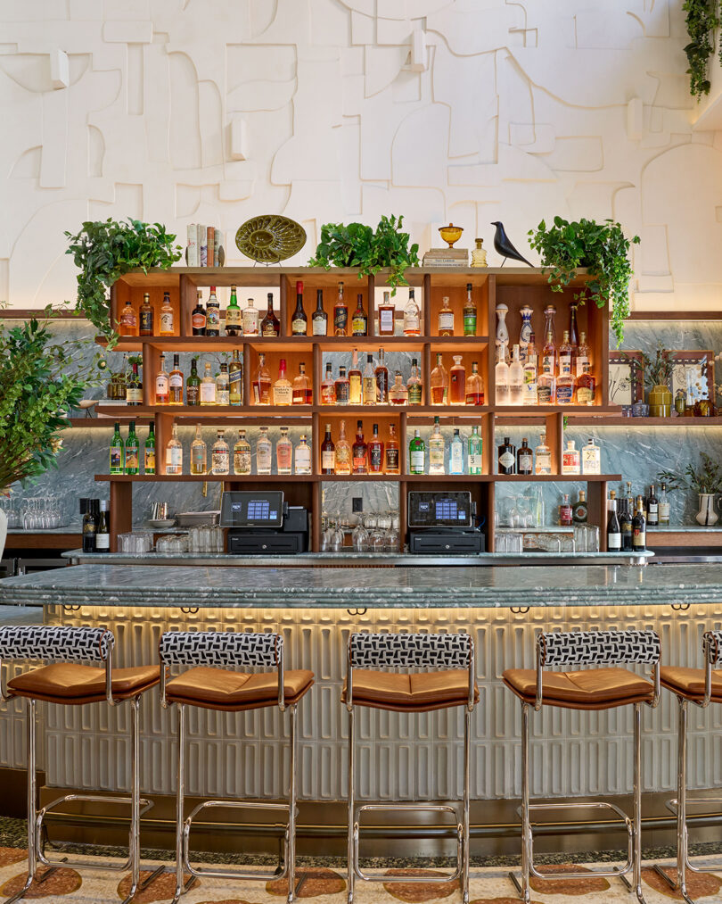 A modern bar by LEGEARD STUDIO with a marble countertop, patterned barstools, shelves stocked with liquor bottles, lush greenery, and decorative wall art.