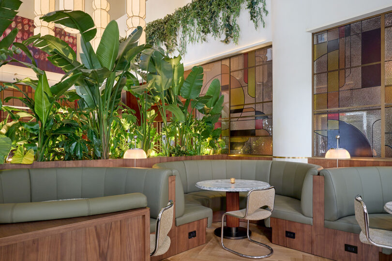A modern restaurant interior by LEGEARD STUDIO with green upholstered booths, marble tables, large indoor plants, and stained glass wall panels.