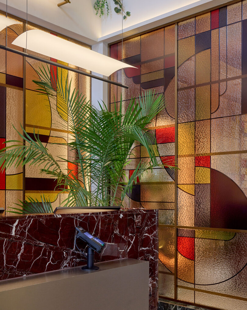 A modern interior by LEGEARD STUDIO features a striking red marble counter, a green potted plant, and geometric stained glass panels in warm tones.
