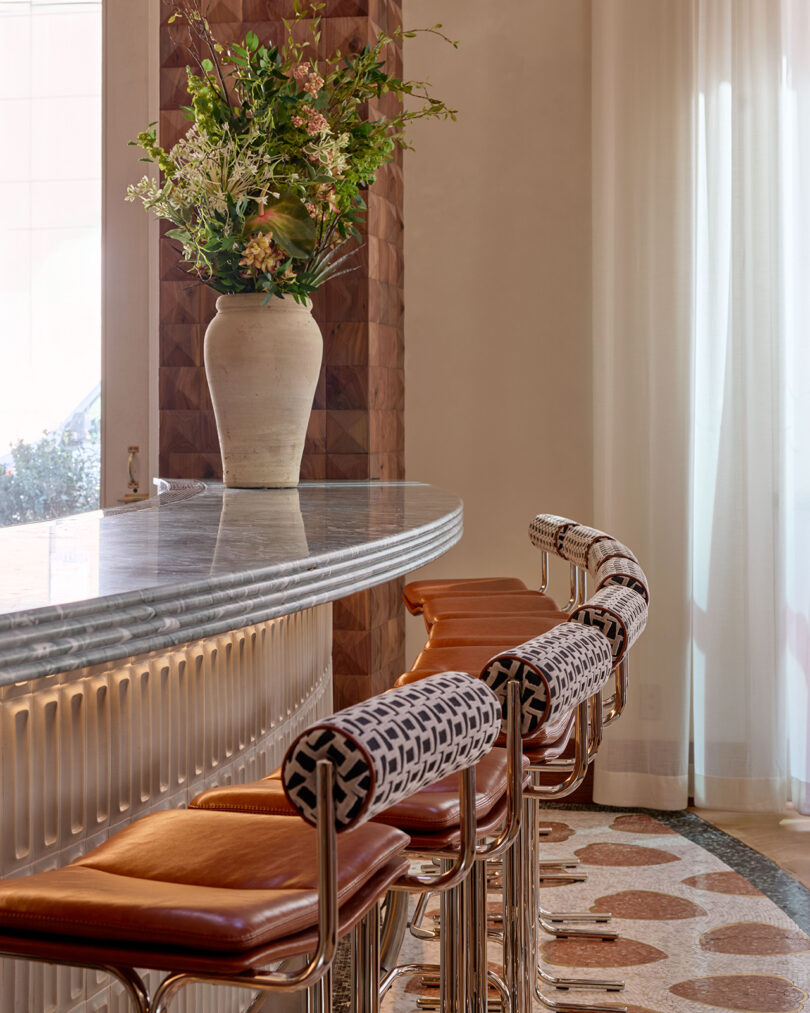 Curved marble bar by LEGEARD STUDIO with patterned barstools featuring leather seats and decorative backs; large vase with flowers sits on the counter.