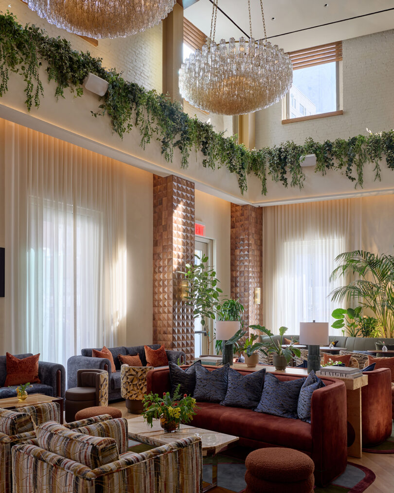 A modern lounge by LEGEARD STUDIO features large windows, lush greenery, plush sofas, armchairs, decorative pillows, and two ornate chandeliers suspended beneath a soaring high ceiling.