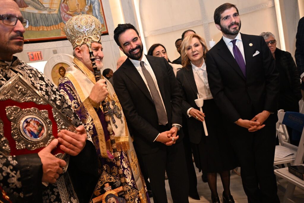A Greek Orthodox Archbishop in ornate gold and purple vestments stands beside a smiling man in a dark suit inside a church.