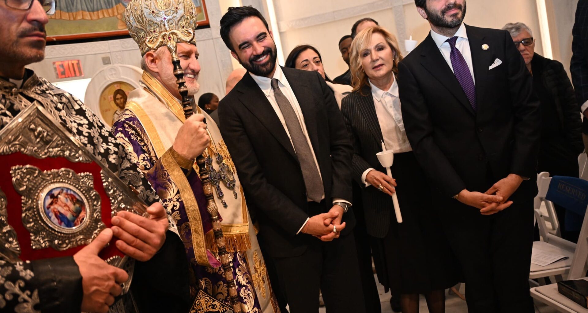 A Greek Orthodox Archbishop in ornate gold and purple vestments stands beside a smiling man in a dark suit inside a church.
