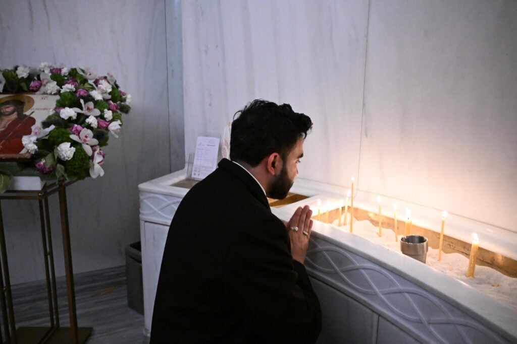 Mamdani kneels in prayer before a row of thin lit candles inside the marble interior of St Nicholas church.