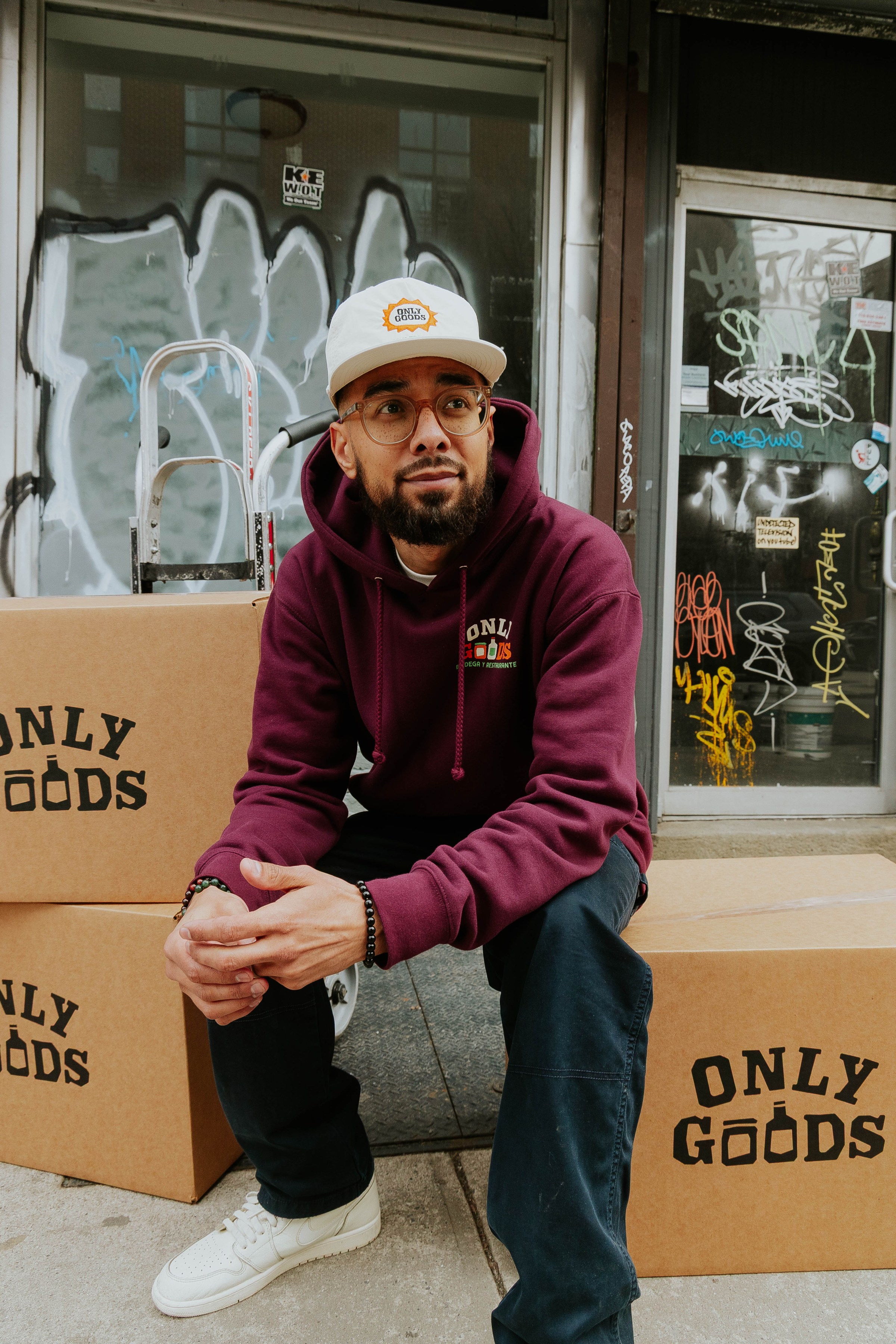 A person sitting on cardboard boxes in front of a shuttered storefront wearing a sweatshirt and cap.