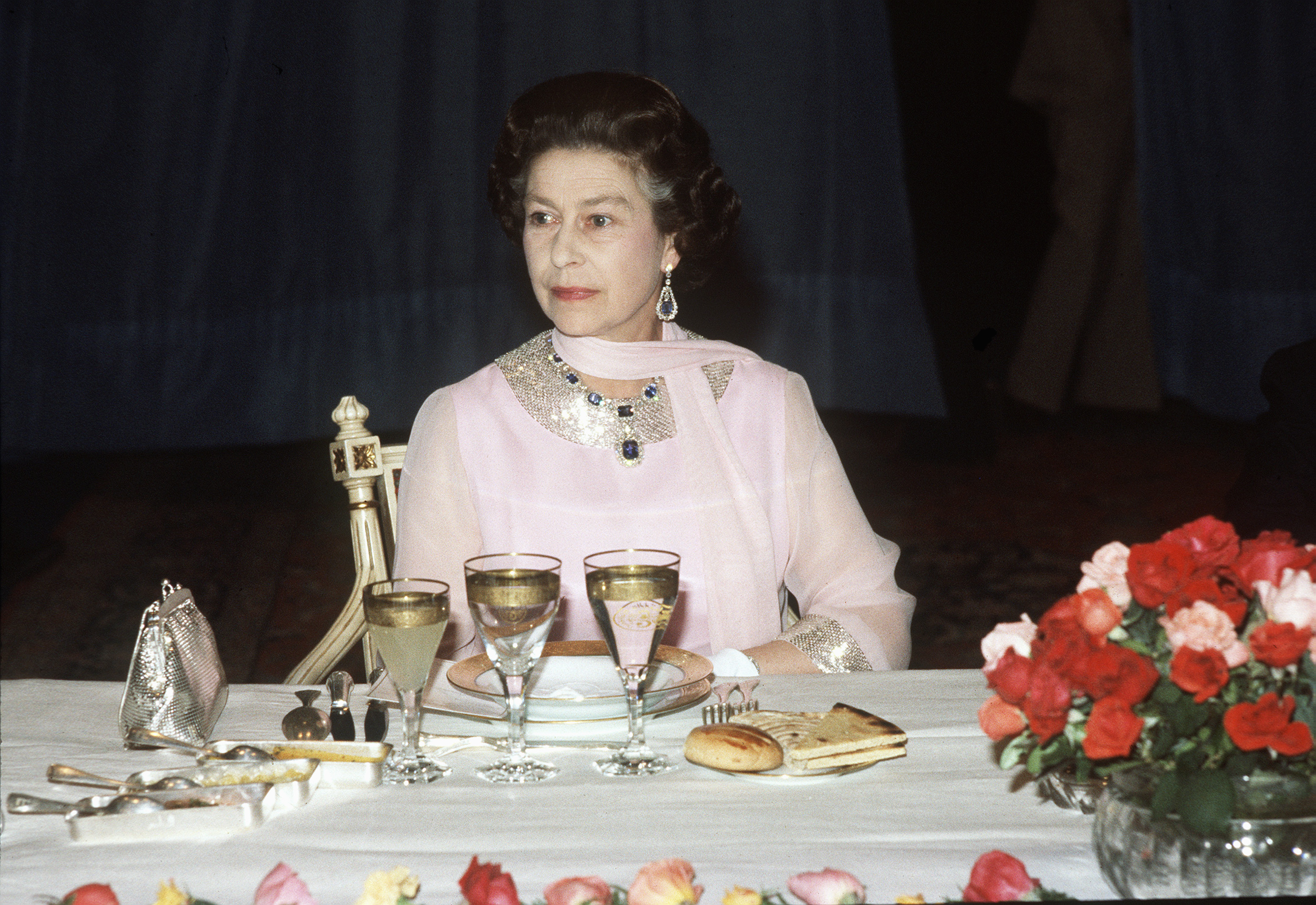Queen Elizabeth wearin ga pink dress and scarf sitting at a dinner table