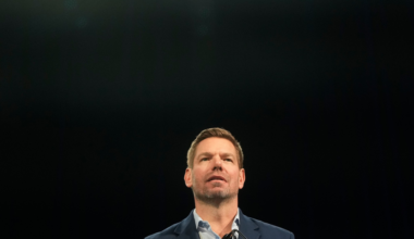 A man with short hair and a trimmed beard stands behind a microphone, wearing a blue blazer and light blue shirt, against a dark background.