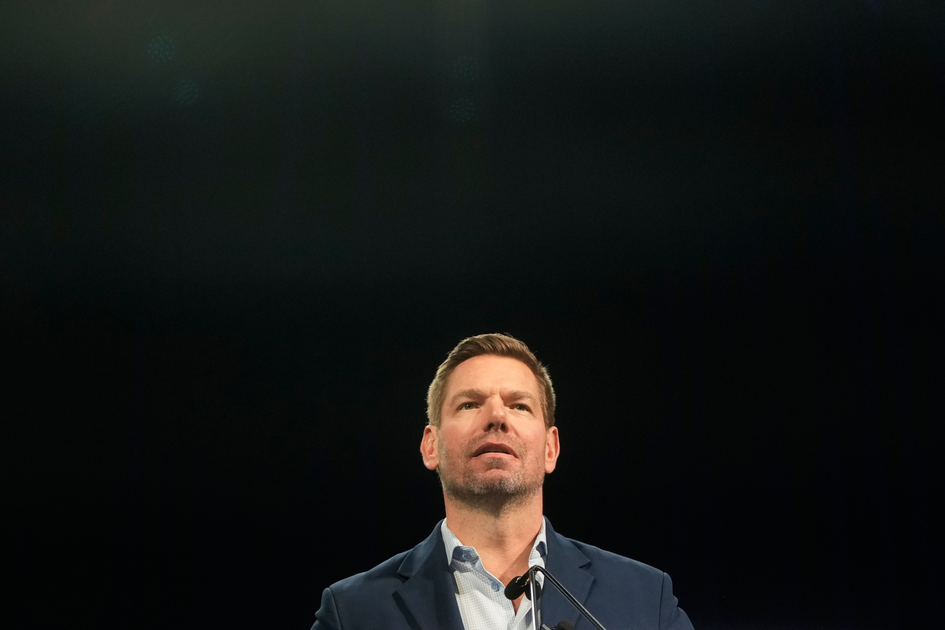 A man with short hair and a trimmed beard stands behind a microphone, wearing a blue blazer and light blue shirt, against a dark background.