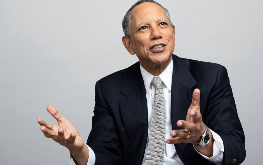 A man in a suit and tie gestures with both hands while speaking, seated against a plain light gray background, reminiscent of a New York Times interview.