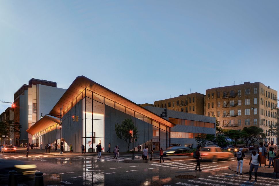 An evening rendering of the renovated Bronx Museum building with a glowing angular glass façade at a busy street corner, illustrating the institution’s upcoming expansion.