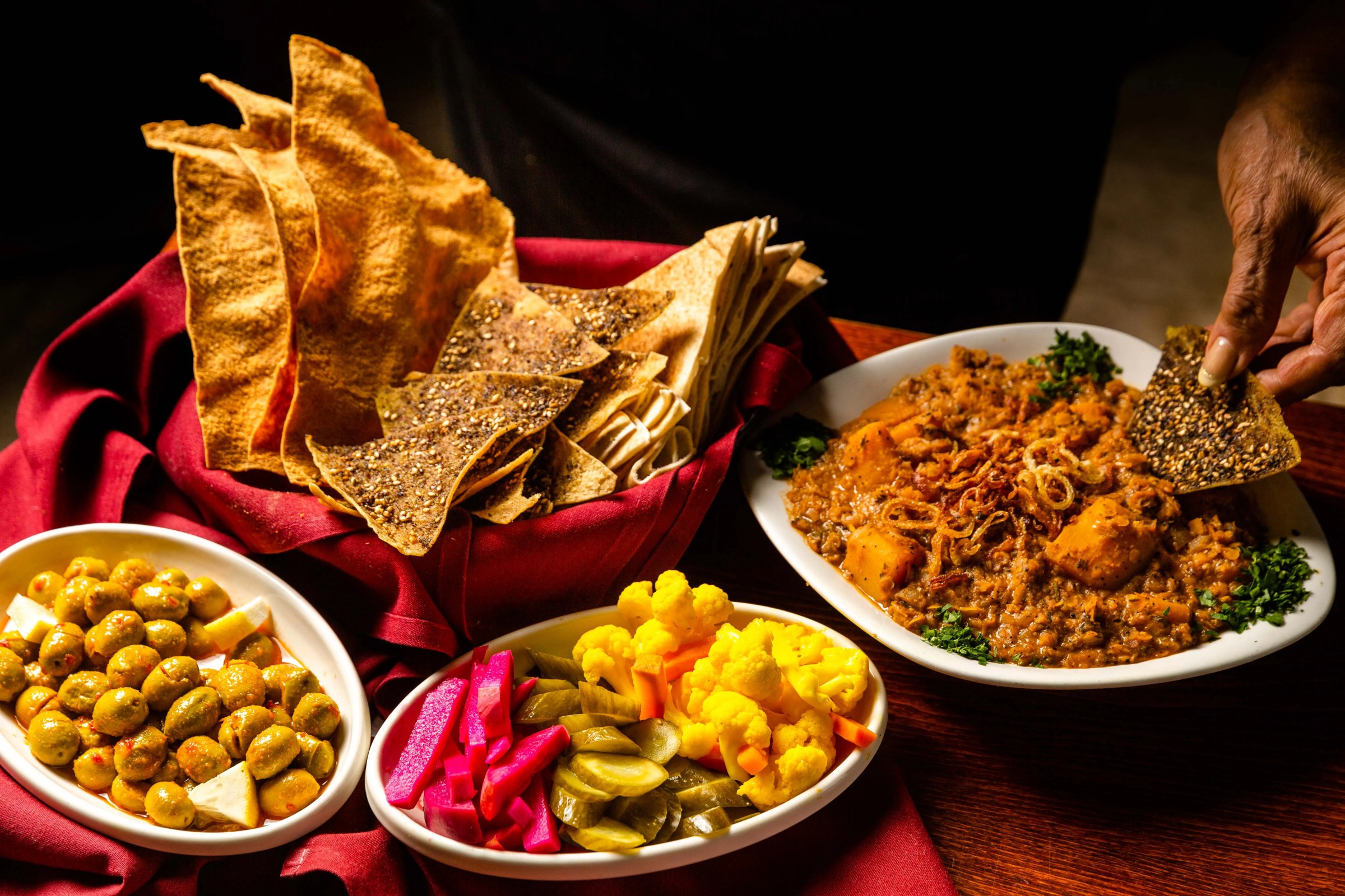 A person takes a scoop of a dip using a seeded pita chip, alongside bright bowls of pickles and olives.