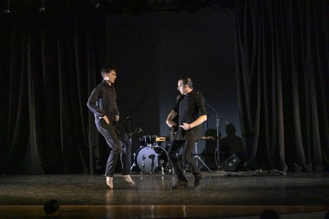 Two male dancers dressed in black face one another with hands resting on the front of the pelvis. Each are jumping up.
