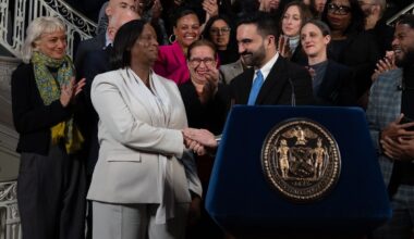 A photo shows a a Black woman wearing a light gray suit shaking the hands of an American man of South Asian descent wearing a black suit and blue tie.  They are standing behind a podium, and a group of people are standing behind them clapping.