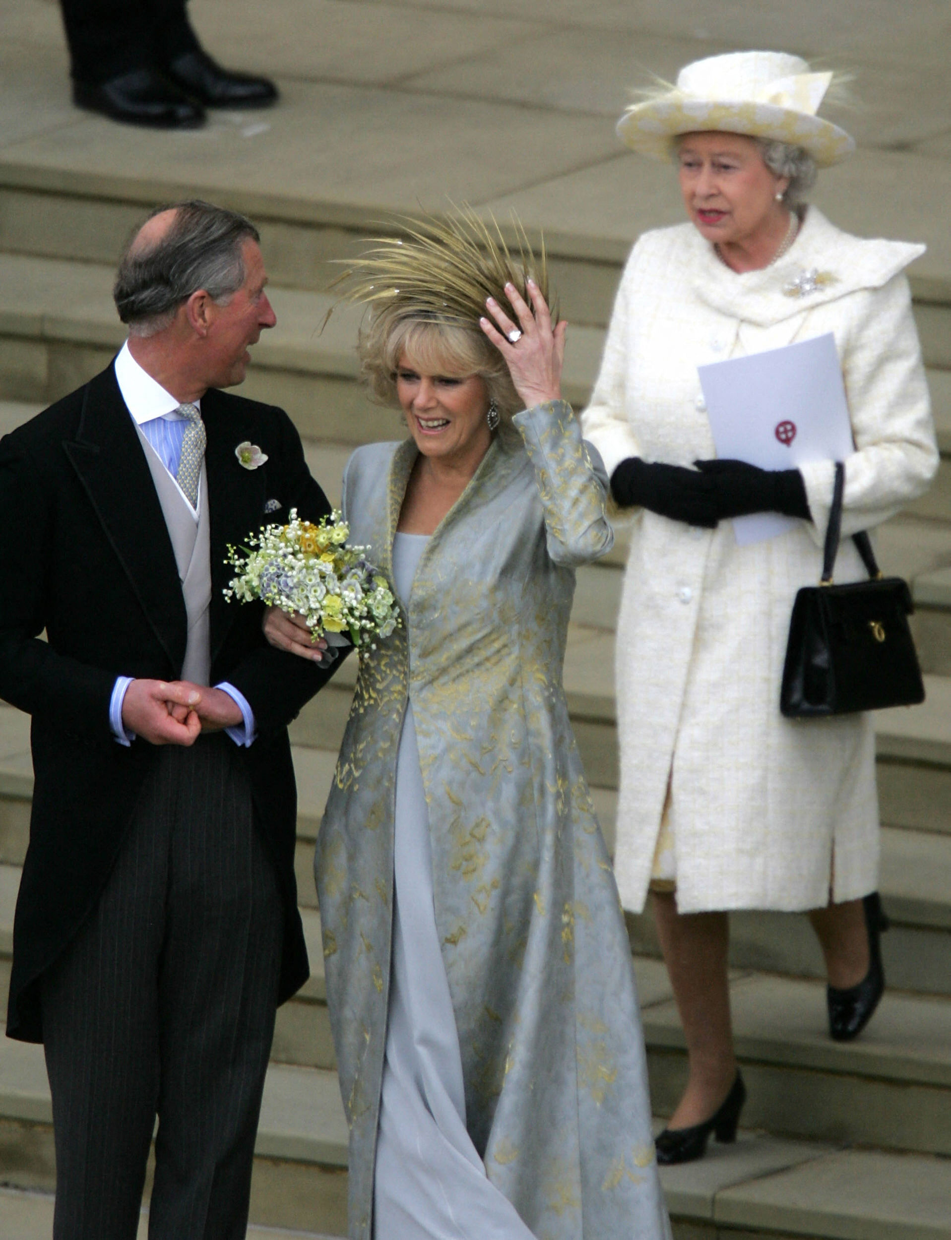 Queen Camilla holding on to her feathered hat on her wedding day next to King Charles and Queen Elizabeth