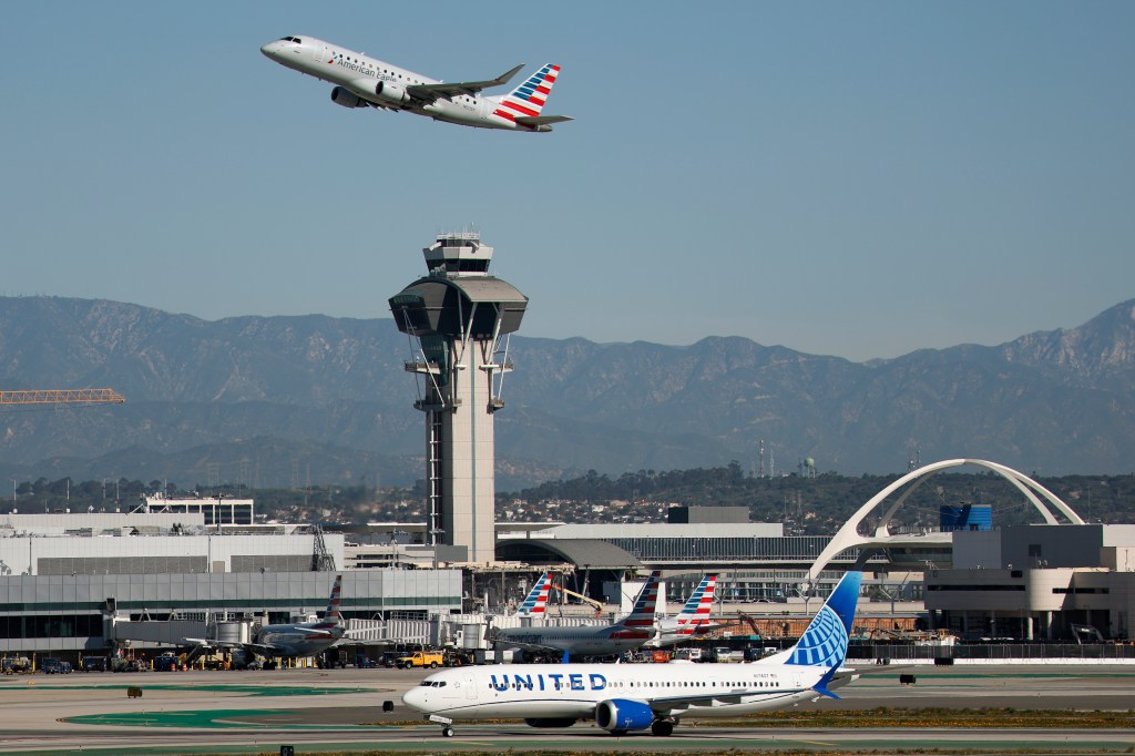 An American Eagle SkyWest Airlines Embraer E175LR airplane departs Los Angeles International Airport as a United Airlines aircraft taxies on the runway, with the air traffic control tower in the background.