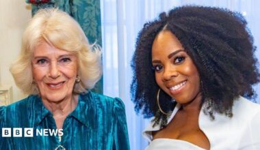 Selina Brown, on the right, has curly black hair to her shoulders and wears a white top while smiling. Queen Camilla, on the left, has shorter blonde hair and smiles, wearing a turquoise dress.
