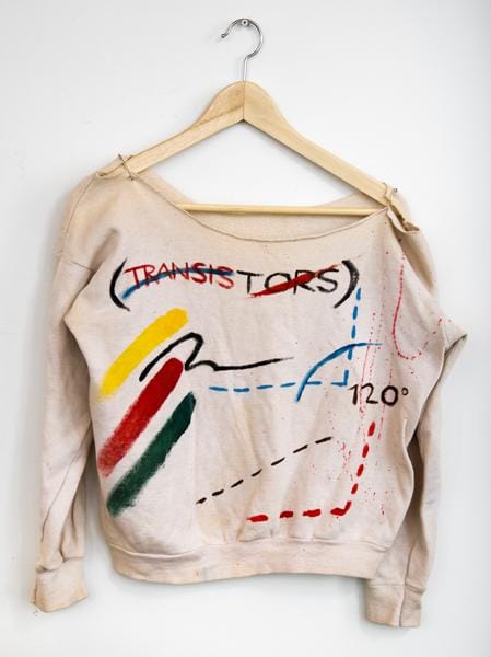 A photograph of a beige sweatshirt featuring abstract decorations including the word 'transitors' rendered in primary hues. The garment is suspended from a wooden hanger against a white wall