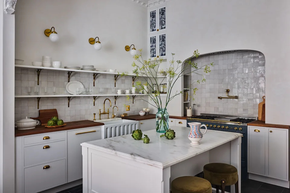 A vintage-inspired Ilve range and the toile fabric panels from Stout Textiles on the cabinets add a touch of navy to the white kitchen. Zellige tiles by Zia Tile line the backsplash, offset by unlacquered brass fixtures, including Rejuvenation hardware, House of Antique Hardware brackets, and a deVOL sink tap and pot filler. “We tried to stick to unlacquered brass for as many of the fixtures as possible, so that it would start to patina and look a little crumbly over time,” says Weed.